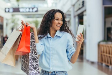 Çekici genç bir Latin Amerikalı kadın alışveriş merkezinde ellerinde renkli poşetlerle alışveriş yapıyor. Telefonu kullanıyor, selfie çekiyor, gülümsüyor..