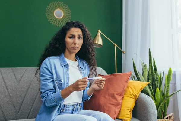 stock image A beautiful young Latin American woman is shocked by the news of an unplanned pregnancy. She is sitting at home on the couch, holding a positive home pregnancy test in her hands. Confused, she looks