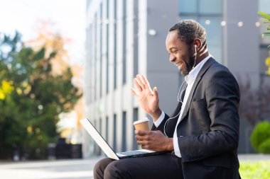 Başarılı ve mutlu bir Afro-Amerikan işadamının video çağrısı, ofis binasının dışında kulaklıklı bir adam iş arkadaşlarıyla ve ortaklarıyla konuşuyor, internette dizüstü bilgisayar kullanıyor, takım elbiseli bir patron..