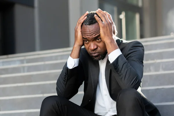 Upset and fired office worker sitting on stairs outside office building close up businessman in business suit depressed lost money bankrupt african american worker sitting sad.