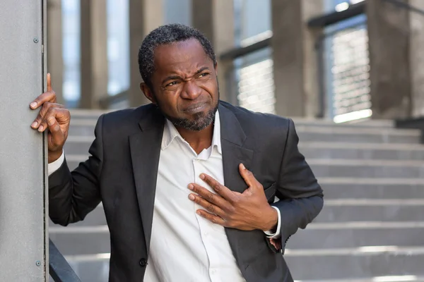 stock image Heart attack , african american businessman has severe chest pain , man holding his hands over heart , mature boss outside office building on stairs.