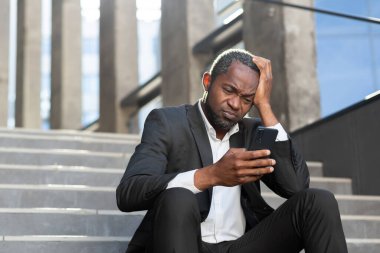 Üzgün Afro-Amerikalı iş adamı depresyonda haberleri okuyor. Ofis çalışanının merdivenlerinde oturuyor. Elinde akıllı telefon var..