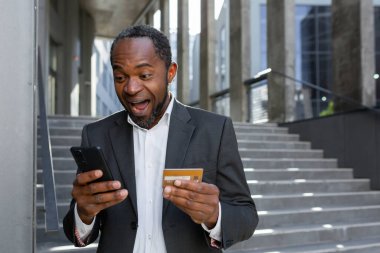 Mutlu Afro-Amerikan iş adamı ofis binasının dışında telefon ve banka kredi kartıyla online alışveriş ve para transferi yapıyor, takım elbiseli olgun patron gülümsüyor..