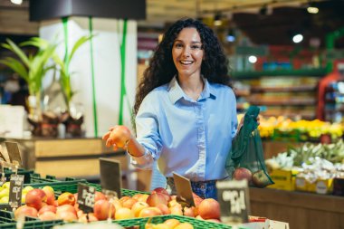 Süpermarkette alışveriş yapan mutlu kadın portresi, İspanyol kadın elmaları ve meyveleri seçiyor, gülümsüyor ve kameraya bakıyor, market sepeti ise malları seçiyor..