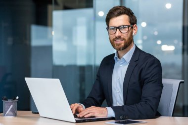 Olgun, başarılı ve mutlu iş adamının portresi, sakallı, gülümseyen ve kameraya bakan son sınıf öğrencisi, ofis içinde iş elbisesi içinde dizüstü bilgisayarla çalışan yatırımcı..