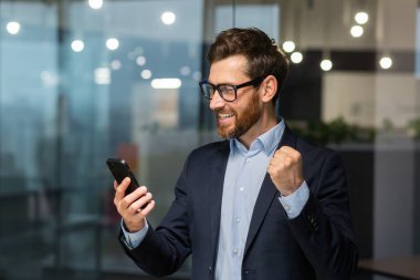 Başarılı bir iş adamı telefon kullanıyor, zaferi ve başarıyı kutlayan bir adam, el ele tutuşup zafer jesti yapıyor, ofis içinde takım elbiseli patron pencerenin yanında duruyor..