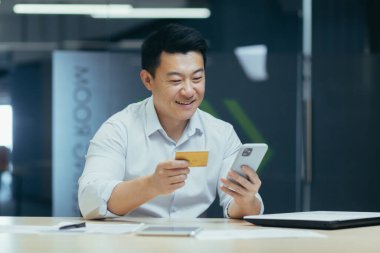 Genç Asyalı adam ofisinde oturuyor, kredi kartıyla online ödeme yapıyor, telefonla sipariş veriyor, mobil bankacılık uygulaması ile işlem yapıyor..