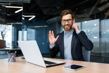 Modern bir ofisteki başarılı bir iş adamının portresi, takım elbiseli bir adam kulaklıkları video görüşmesi ve uzaktan iletişim için kullanır, bir yatırımcı kameraya bakar ve dizüstü bilgisayar kullanır..