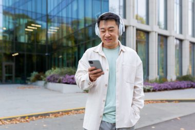 Sıradan kıyafetler içinde Asyalı bir adam kulaklıkla müzik dinliyor gülümsüyor ve mutlu, akıllı telefon uygulaması kullanıyor podcast ve online radyo dinliyor, modern ofis binasının dışında bir adam.