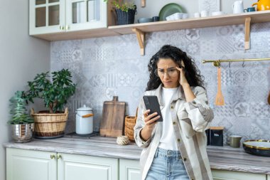 Şok olmuş Latin Amerikalı kadın mutfakta elinde telefonla evde dikiliyor. Üzgün, telefonuna bakıyor, beklenmedik ve kötü mesajlar alıyor, haberler....