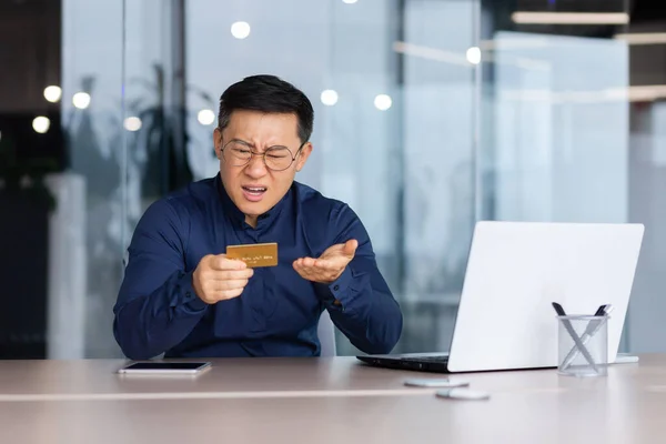 灰心丧气的亚洲人试图在网上商店里进行银行业务和购买 商人在办公室里拿着笔记本电脑 手里拿着银行卡 手忙脚乱地工作 — 图库照片