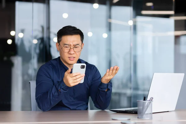 Businessman reading bad news online from phone, asian man disappointed and sad looking at smartphone screen, man in shirt working inside office using laptop at work.