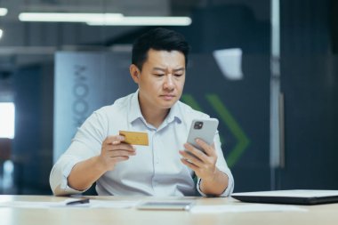 Ciddi bir Asyalı genç adamın elinde kredi kartı ve telefon var. Parasal işlemler yapıyor, online alışveriş yapıyor, hesabı kontrol ediyor. Ofisteki masada oturuyor..
