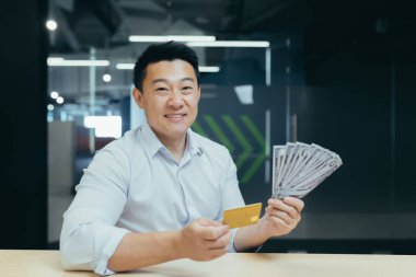 Mutlu genç bir adam elinde kredi kartı ve nakit para tutuyor, kredi kartı, kredi kartı, yatırım alıyor. Ofiste oturmuş, kameraya gülümsüyorsun..