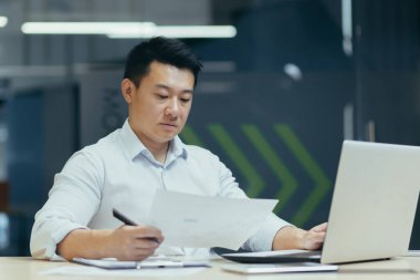 Genç ciddi bir adam, Asyalı bir avukat, masanın başında oturuyor, elinde belgeler ve anlaşmalar var. Dizüstü bilgisayarda konsantre çalışma.