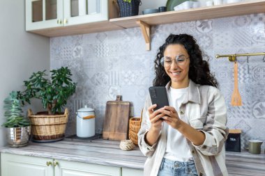 Genç İspanyol kadın mutfakta telefonuyla bekliyor. Çevrimiçi alımlar, siparişler, ev teslimatı yapıyor..