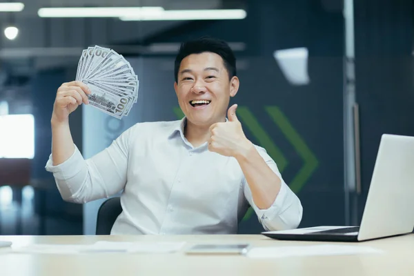一个快乐的亚洲年轻人 拿着笔记本电脑坐在办公室的桌子旁 手里拿着一把钱 手里拿着现金 伸出了一个大指头 微笑的男人看着镜头 指着镜头 — 图库照片