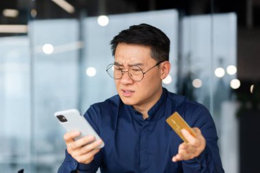 Aldatılmış online alıcı, Asyalı işadamı banka işlemleri ve çevrimiçi alışveriş yapmaya çalışıyor, banka kartı ve telefonu elinde tutan mutsuz bir adam..