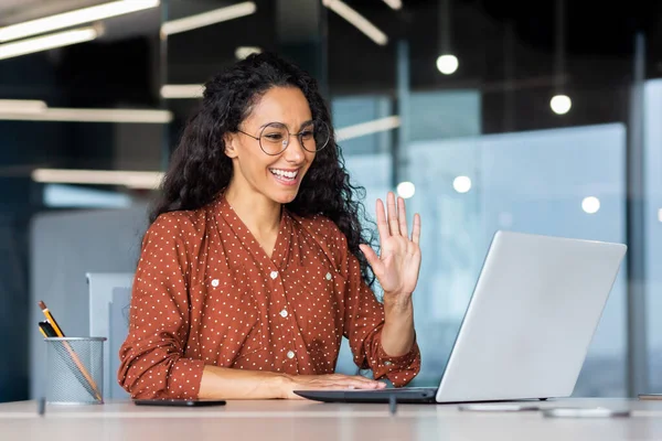 Stock image Successful and beautiful hispanic woman working inside modern office building, businesswoman using laptop for video call smiling and waving, greeting gesture, online conference with colleagues.