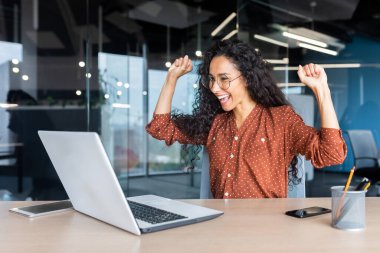 İspanyol iş kadını zafer başarısını kutluyor, ofis içinde kıvırcık saçlı çalışan iyi haberler okuyor, iş yerinde bilgisayarını kullanıyor, el ele tutuşuyor ve mutlu zafer jestleri yapıyor..