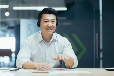 Çevrimiçi iş eğitimi. Genç gülümseyen Asyalı adam ofiste kameranın önünde, kulaklıkla oturup konuşuyor, online bir toplantı düzenliyor,