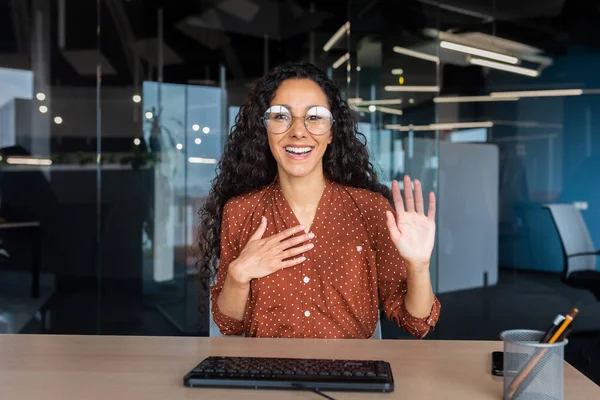Online customer support video call, Latin American woman looking at web camera smiling and consulting customers, helpline worker working inside office.
