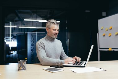 Modern ofiste çalışan olgun, başarılı bir patron. Üst düzey, gri saçlı iş adamı. Dizüstü bilgisayarla çalışıyor..