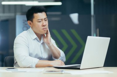 Dişim ağrıyor. Genç Asyalı bir adam masadaki ofiste oturuyor, dizüstü bilgisayarda çalışıyor, yanağını tutuyor, diş ağrısı çekiyor..