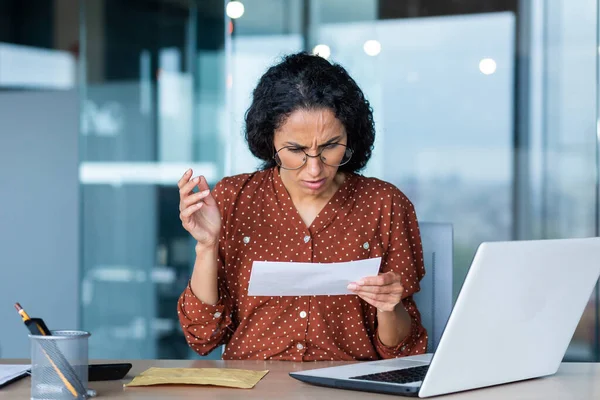 A young Latin American woman, secretary, manager, accountant is worriedly holding a letter, document, account. She sits confused in the office at the desk.