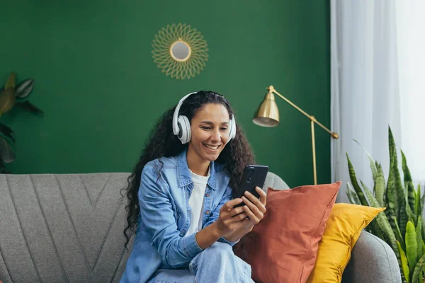 Young smiling hispanic woman at home with headphones and phone listening to music online using smartphone app sitting on sofa in living room and dancing.