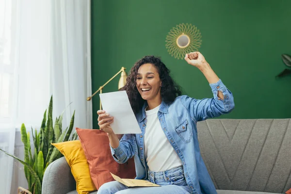 Happy woman at home received nice notification letter reading and happy smiling, Hispanic woman at home with holding message envelope sitting on sofa in living room.
