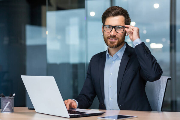 Portrait of a successful young businessman, lawyer, legal defender. Confidently looks into the camera, holds glasses, laughs, sits in the office at the table, works at the laptop.