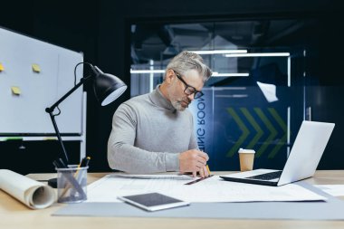 Yakışıklı, gri saçlı mimar, tasarımcı, mühendis, kağıt üzerinde kalemle bir çizim yapıyor, bir plan yapıyor, ofisteki masada projeye konsantre oluyor..