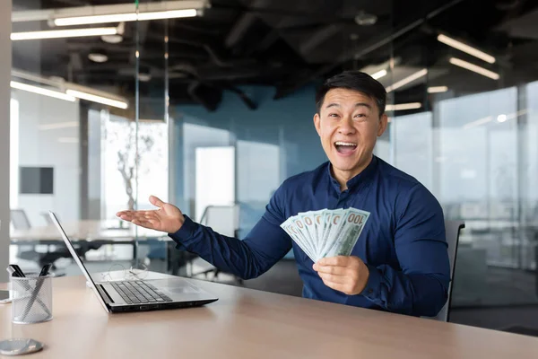 Happy asian banker investor working inside modern office building, man looking at camera and happy showing money cash dollars deal income.
