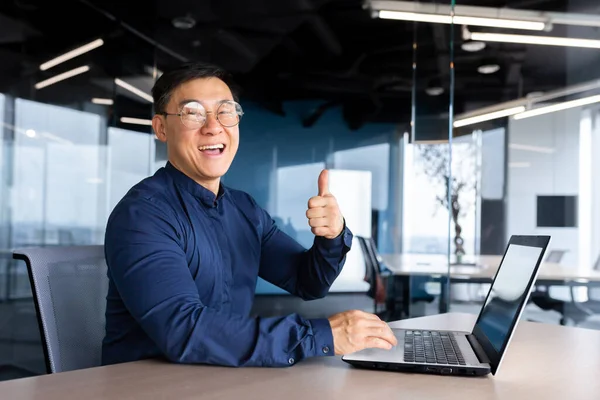 Happy and satisfied asian programmer looking at camera and smiling holding thumb up, businessman working inside office using laptop at work.