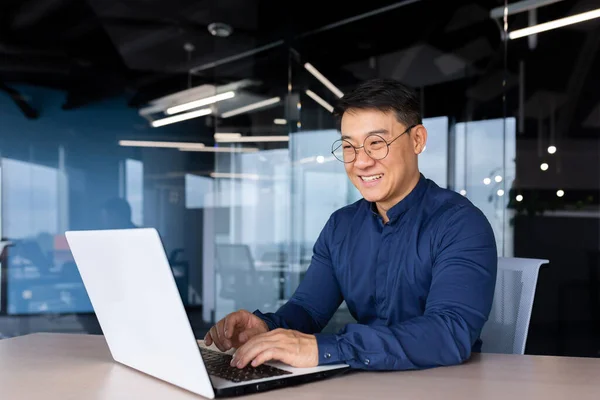 Cheerful and smiling Asian man working inside office, businessman in shirt and glasses using laptop, engineer developer programmer typing code programming software.