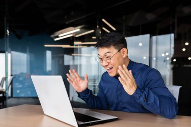 Başarılı Asyalı adam zaferi ve başarıyı online olarak kutluyor. İş adamı dizüstü bilgisayara bakıyor ve zafer jesti yapıyor..