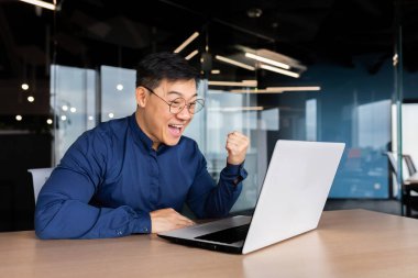 Başarılı Asyalı adam zaferi ve başarıyı online olarak kutluyor. İş adamı dizüstü bilgisayara bakıyor ve zafer jesti yapıyor..