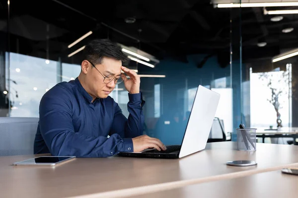 Asian boss working inside office, man overtired with headache, businessman in shirt and glasses using laptop at work, sitting at desk at workplace.