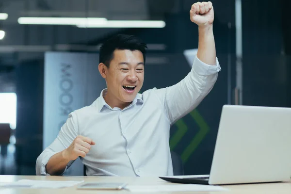 Young happy Asian man at the desk in the office celebrating success, victory, good news, deal. Sitting at the table, looking at the laptop, raising his hands, dancing.