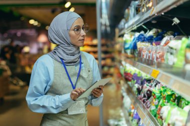 Markette kadın satıcı, tabletle tesettüre girdi, ürünleri kontrol etti, cep bilgisayarı kullandı, Müslüman kadın, ürün ve mallarla rafların yanında..