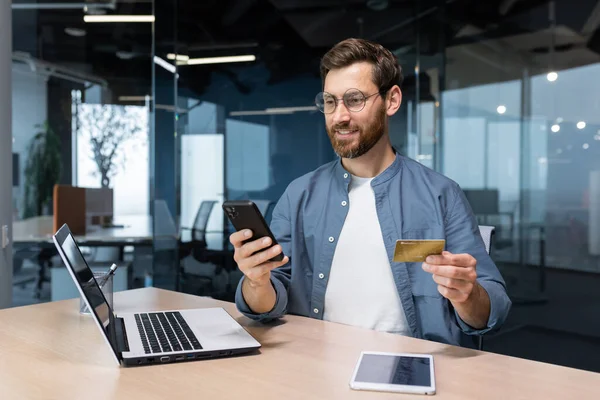 Cheerful and smiling bearded businessman doing online shopping and bank money transfer, freelancer holding bank credit card and smartphone, man sitting at desk inside office.