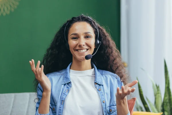 Joyful hispanic woman with video call headset at home smiling and looking at camera, woman studying and working online remotely sitting on couch at home.