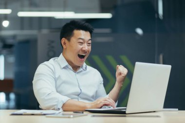 Genç iş adamı serbest çalışan Asyalı ofisteki laptop monitörüne bakıyor, önemli iyi haberler alıyor, seviniyor, eliyle puan kazanıyor evet, mutlulukla bağırıyor..