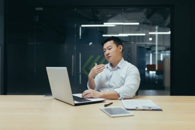 Genç bir Asyalı adam masanın ofisinde oturuyor, bir dizüstü bilgisayar üzerinde çalışıyor. Elini yüzüne doğru sallıyor. Kendini kötü hissediyor, sıcak hissediyor, temiz havaya, tıbbi yardıma ihtiyacı var..