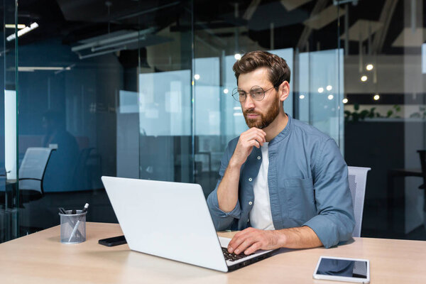 .Serious pensive businessman in shirt thinking about decision sitting at table in modern office, man with beard is using laptop at work.