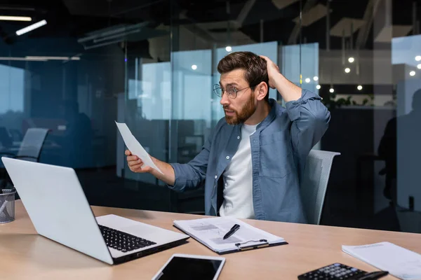 Problems at work. Worried and nervous young male businessman, freelancer shocked, holding documents, bills in his hands. holding his head with his hand. Sitting in the office at a table with a laptop.