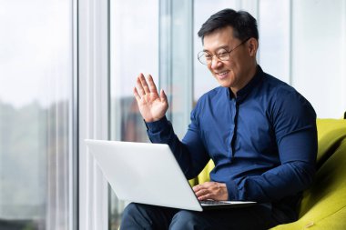 Genç bir Asyalı erkek öğrenci kampüste oturuyor, internetten bilgisayarla iş birliği yapıyor. Video aracılığıyla konuşuyor, elleriyle selamlıyor, gülümsüyor..