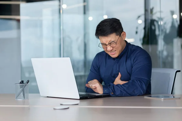 Heart attack at work. A young Asian man is sitting at a desk in the office, holding his heart. A stroke, holding his chest. grimaced, feels severe pain, needs help.