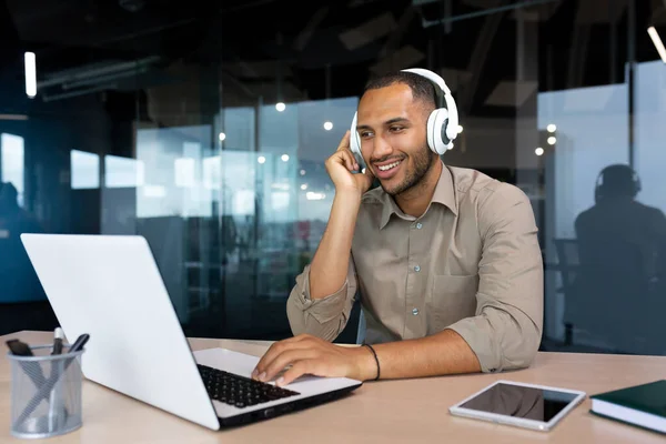 Young hispanic businessman in shirt working inside office using laptop at work, man with headphones listening to music and audio podcasts at work.
